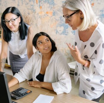 three women stand and sit around a computer 