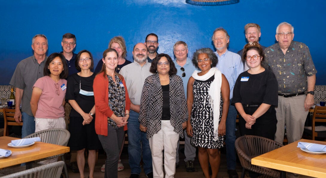 a group of about a dozen diverse individuals stands in front of a blue wall in a restaurant; tables are in the foreground